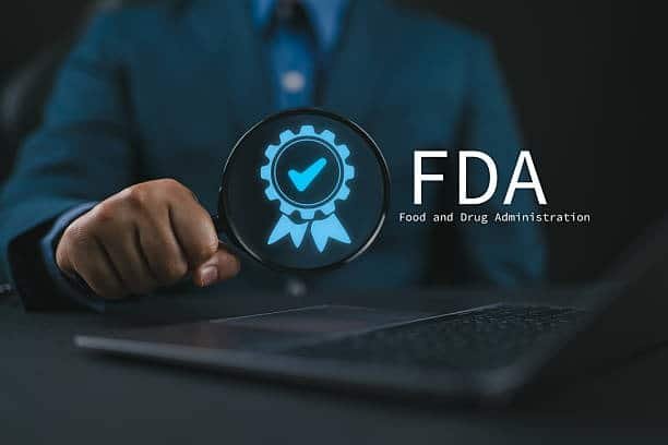 Alt text: Business professional holding a magnifying glass with a blue approval badge beside the FDA (Food and Drug Administration) logo, representing regulatory compliance and product approval.