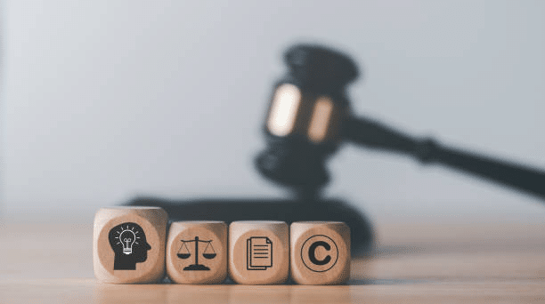 Wooden blocks featuring icons for innovation, legal justice, document protection, and copyright, with a judge’s gavel in the background, representing intellectual property law